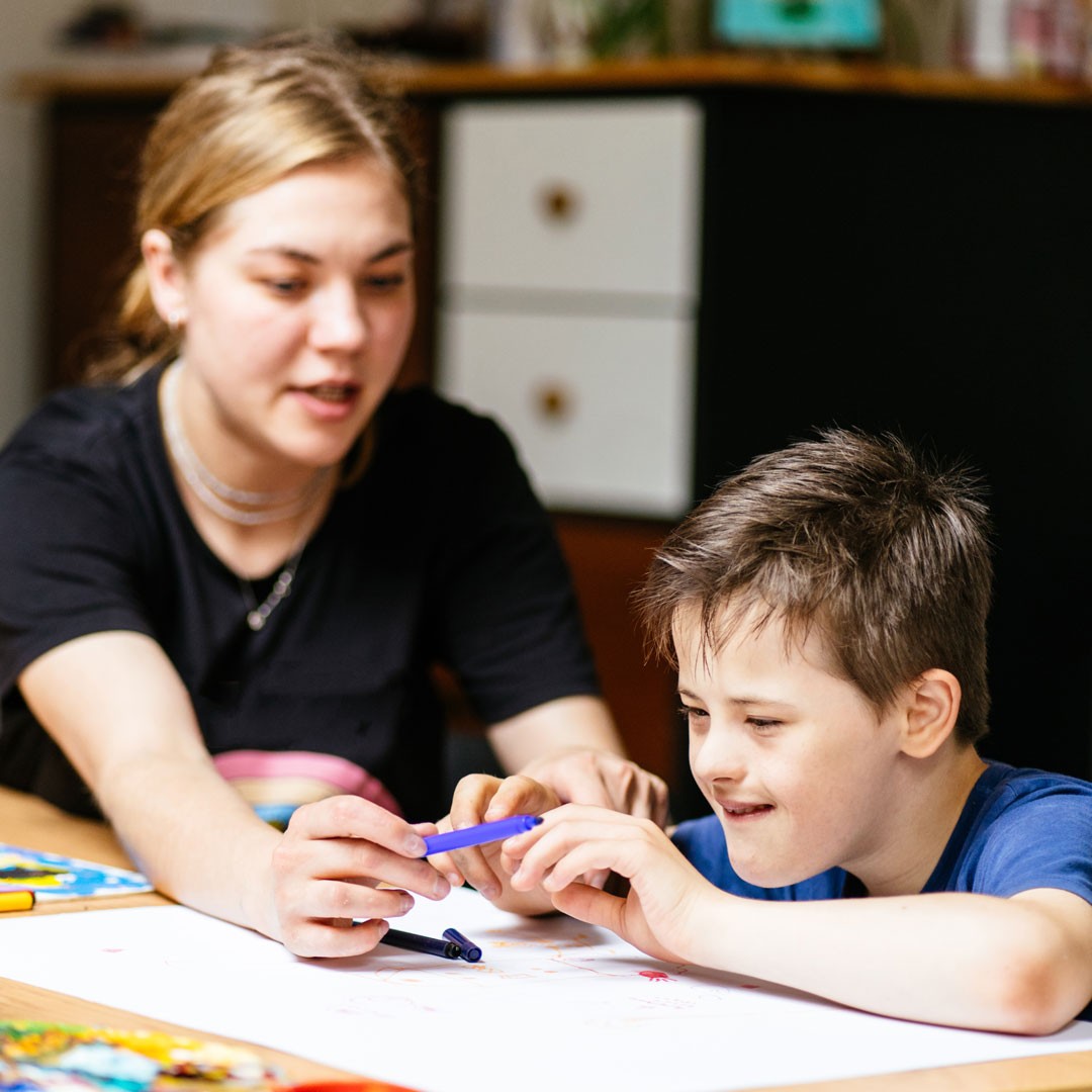 Close up of young female teacher sitting at desk with a Down syndrome schoolboy. Color painting on the paper for disabled kids, autism childs who are down syndrome and student teacher.
